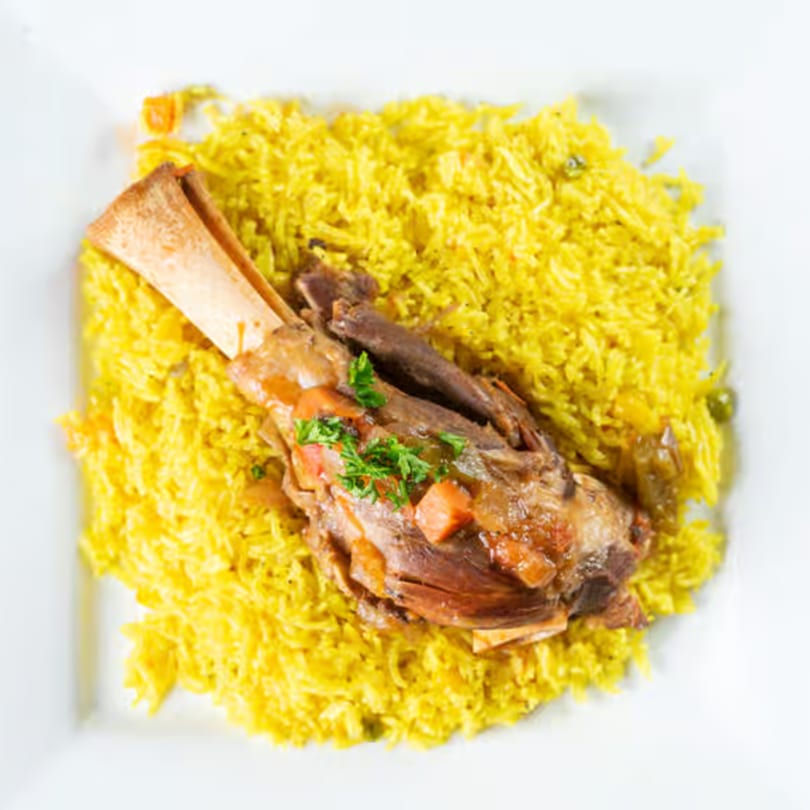 Braised lamb shank with vegetables and parsley, served over a bed of yellow rice.